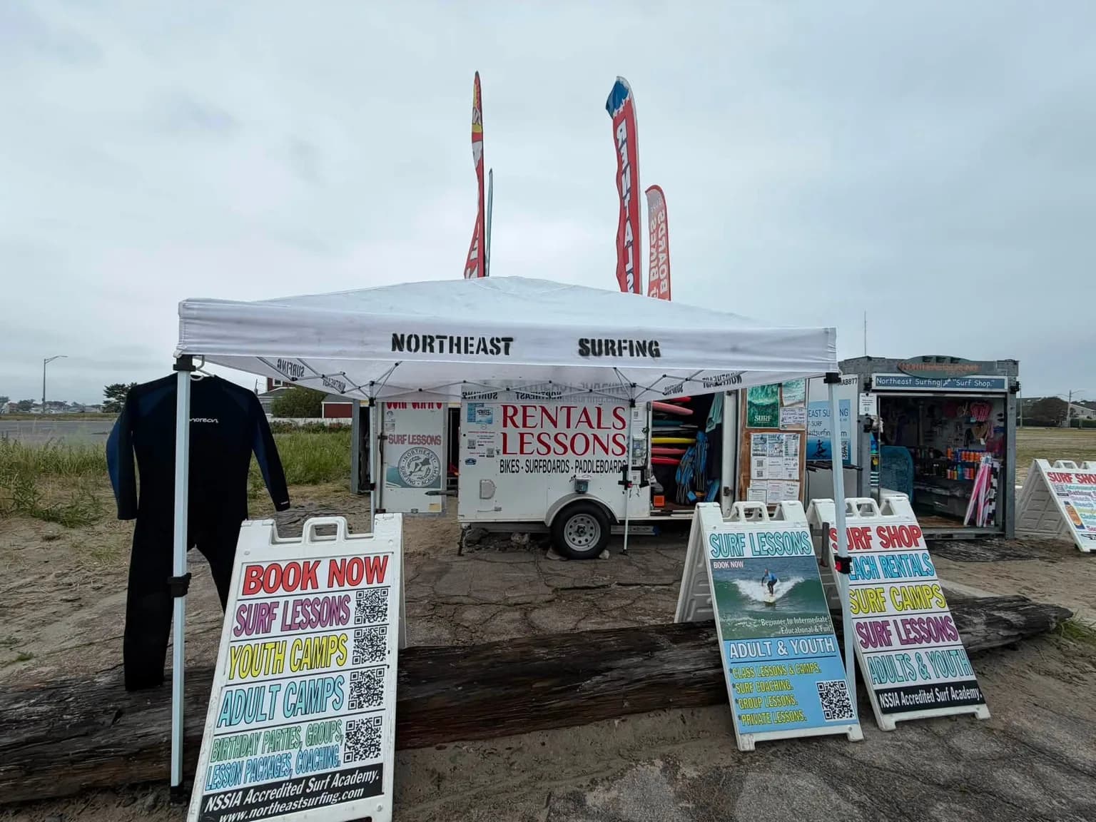 Northeast Surfing's stand on Nantasket Beach: a branded canopy, a rental trailer and a surf shop, with boards advertising lessons and camps