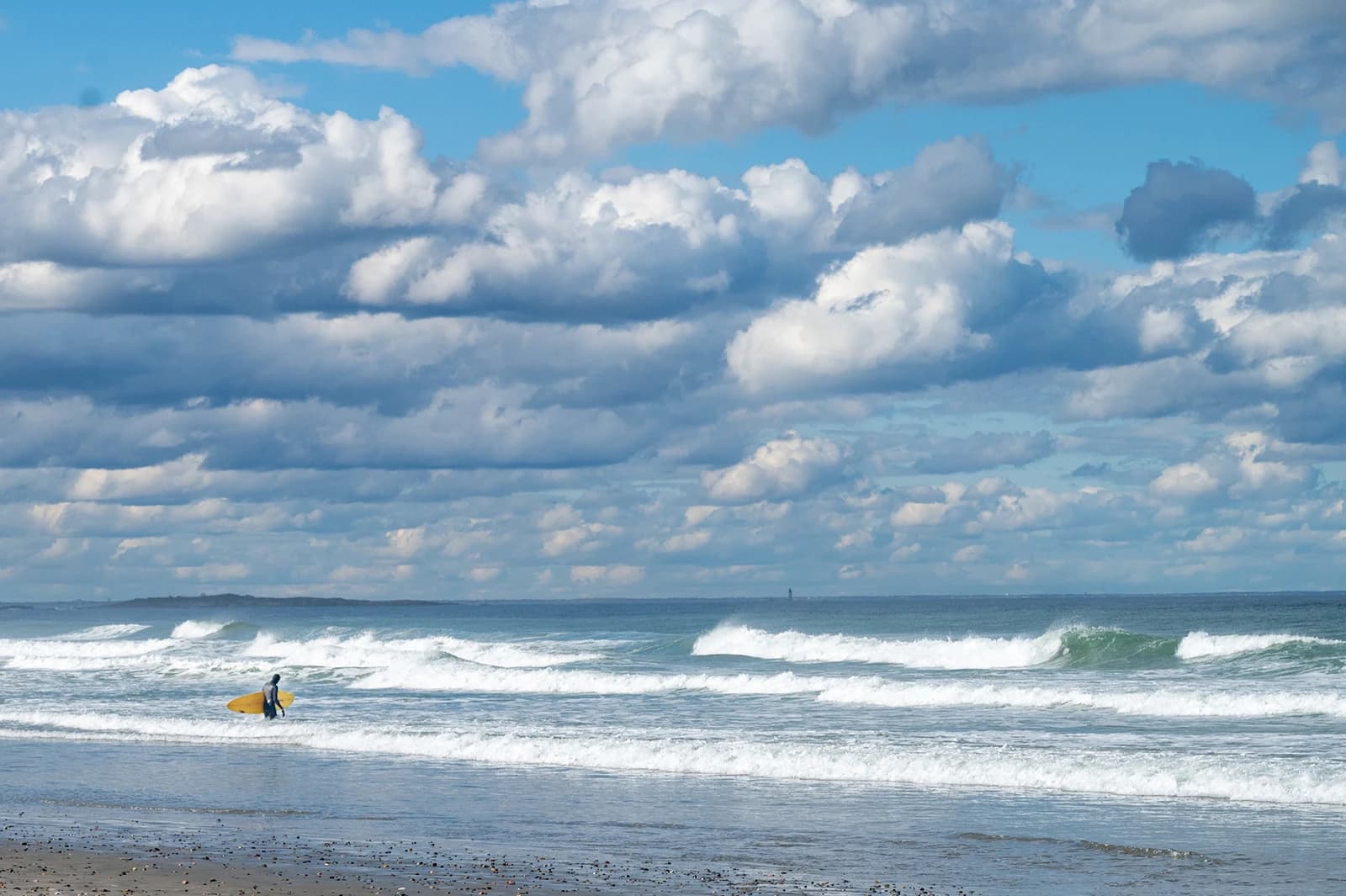 A surfer carrying a yellow board into the breaking waves at Nantasket Beach under a big cloudy sky